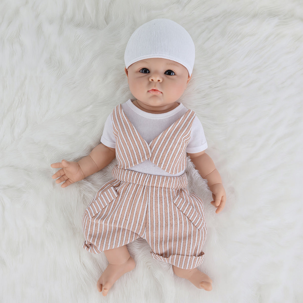 A realistic baby doll with a white knit cap, lying on a white faux fur surface. The doll is dressed in a white short-sleeve shirt and matching white and beige vertically striped overalls.