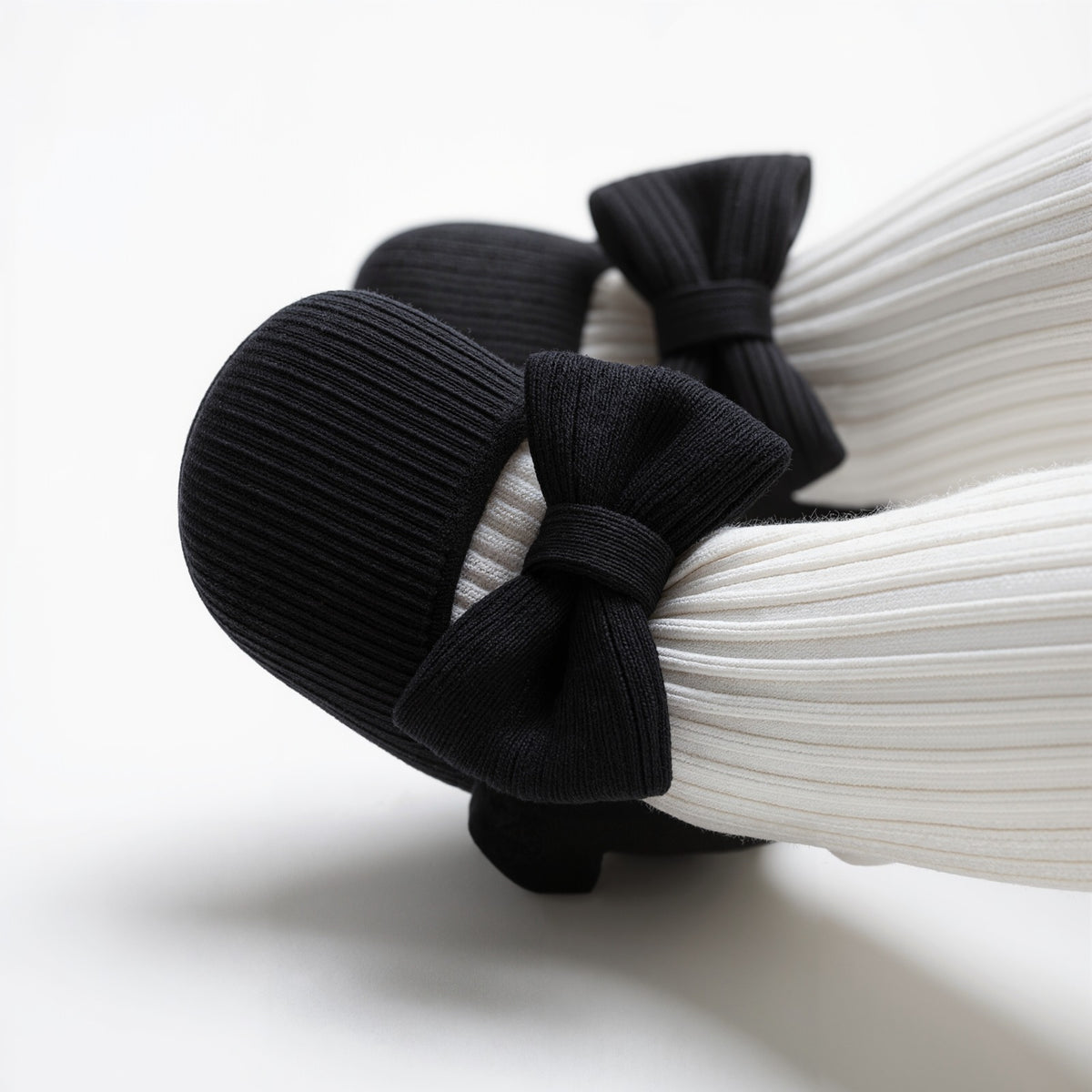 A close-up of a pair of black ribbed flat shoes with large bow details, displayed on white pleated mannequin legs against a clean white background.