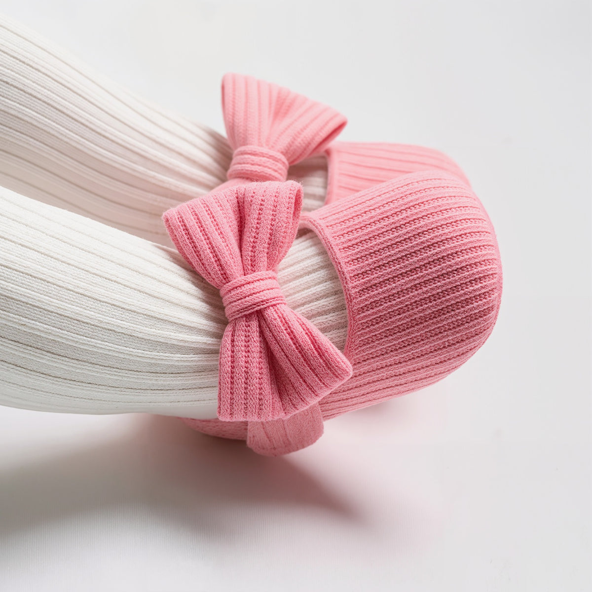 A close-up of a baby's feet wearing white ribbed tights and pink ribbed booties with matching pink bows, set against a clean white background.