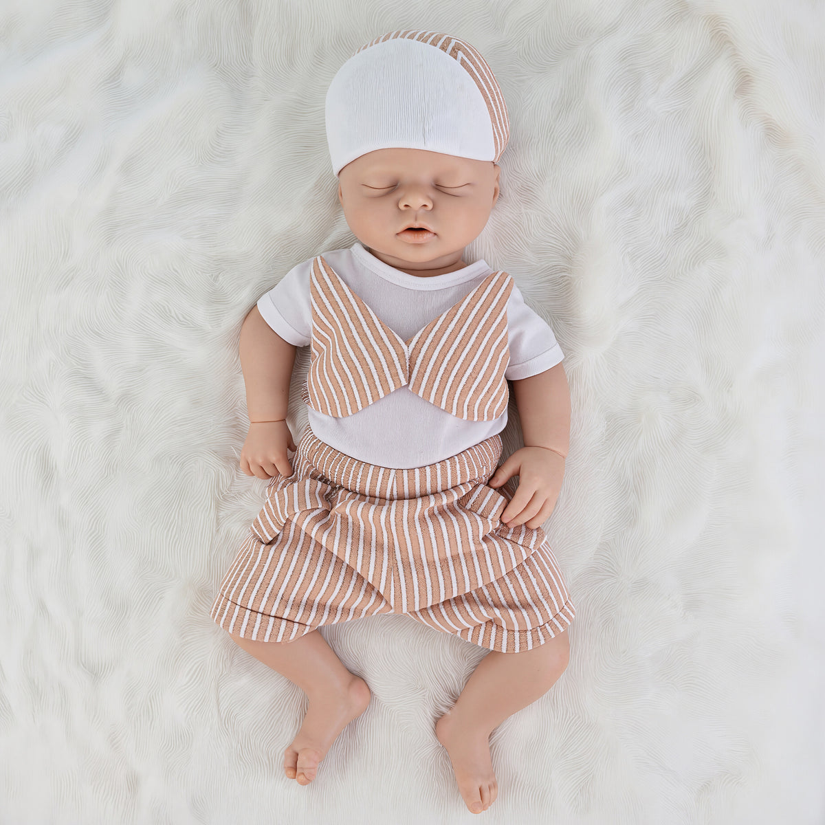 A realistic sleeping baby doll wearing a white short-sleeve shirt, peach and white striped bow tie, matching striped shorts, and a striped beanie, lying on a white faux fur background.