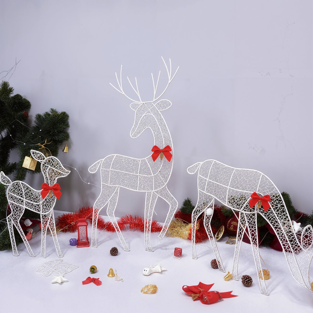 Three white wire-frame reindeer decorations with red bows are arranged on a white surface with holiday ornaments, pinecones, and a Christmas tree in the background.